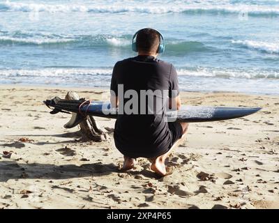 Mann mit Kopfhörern und schwarzem Surfbrett, der auf Sand sitzt und auf das Meer blickt. Rückansicht. Stockfoto