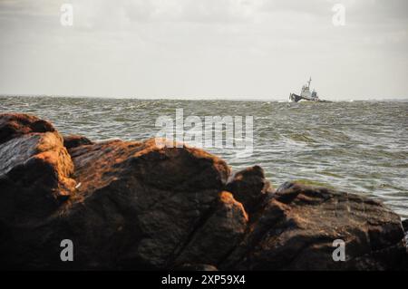 Fischerboot im abgehackten Meer mit felsigem Ufer im Vordergrund unter bewölktem Himmel Stockfoto