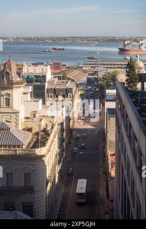 Aus der Vogelperspektive auf eine belebte Straße in Montevideo mit historischen Gebäuden und weit entfernten Schiffen im Hafen. Stockfoto