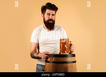 Bärtiger betrunkener Mann mit Bierkrug auf Holzfass. Oktoberfest. Männlicher Brauer mit einem Glas Craft-Bier. Deutsche Traditionen. Gutaussehende Guy-Verkostung Stockfoto