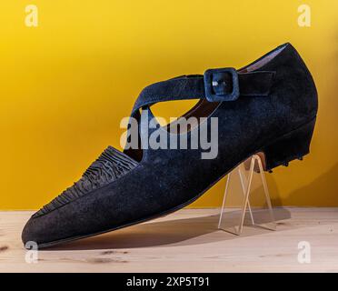 Ein einzelner schwarzer Wildlederschuh mit Riemen und Schnalle, der auf einer Holzoberfläche vor gelbem Hintergrund ausgestellt ist und sein einzigartiges Design und seine Handwerkskunst unterstreicht. Schlüsselwörter: Stockfoto