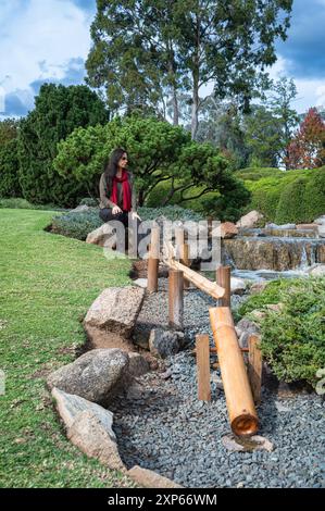 Niedriger Blick auf ein Bambus-Wasserspiel, das zu einer weiblichen Touristen führt, in Reflexion, auf einem Felsen in der Nähe eines kleinen Wasserfalls in einem Japanischen Garten sitzt. Stockfoto