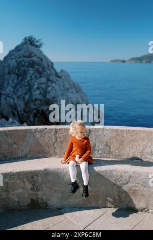 Das kleine Mädchen sitzt auf einer Steinbank auf einer Aussichtsplattform am Meer und schaut weg Stockfoto