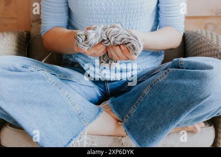 Frau in Jeans, die einen Ball aus grauem Garn hält. Mädchen, das im Sessel sitzt, mit gewebten Fäden in den Händen. Strickkonzept. Kreativer Lebensstil. Strickkunst. Stockfoto