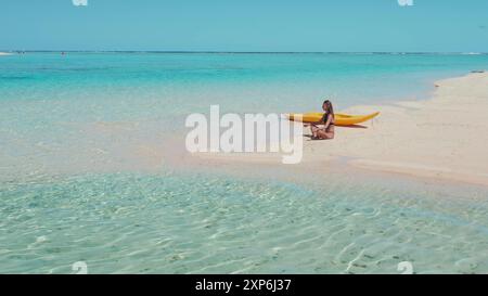 Eine junge Frau übt Yoga auf einer Sandbank auf den malediven mit einem Kajak neben ihr. Das Wasser ist kristallklar und der Sand weiß Stockfoto