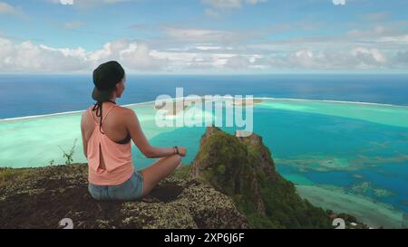 Die junge Frau entspannt sich und meditiert an einer Klippe und genießt den atemberaubenden Panoramablick auf die türkisfarbene Lagune und die üppigen Inseln von maupiti in französisch-polynesien Stockfoto
