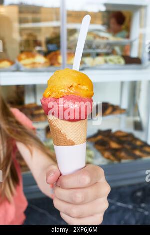 Kegel mit zwei Kugeln Erdbeeren- und Mangofeis, rot und gelb, in der Hand eines Mädchens mit dem Fenster des Süßwarengeschäfts dahinter Stockfoto