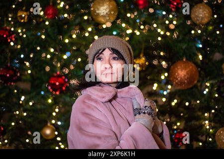 Russische 48-jährige Frau in rosa Mantel, die vor Neujahr vor weihnachtsbaum und Lichtern steht Stockfoto