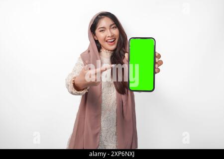 Aufgeregte junge asiatische muslimische Frau im Kopftuchschleier-Hijab, die auf den Kopierraum am leeren Bildschirm des Smartphones zeigt, isoliert auf weißem Hintergrund Studio. R Stockfoto