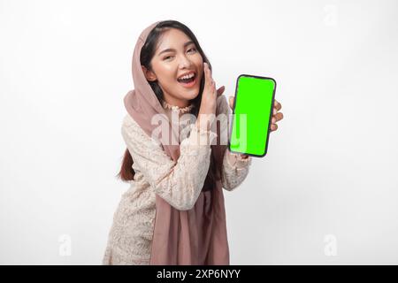 Porträt einer überraschten jungen asiatischen muslimischen Frau im Schleier-Hijab mit leerem Bildschirm auf dem Telefon mit Kopierraum im Inneren, während sie eine Flüsterhand-Geste macht. Stockfoto