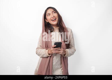 Nachdenkliche asiatische muslimische Frau, die ein Smartphone hält, während sie sich fragt und denkt, mit Kopierraum darüber, isoliert durch weißen Hintergrund Stockfoto