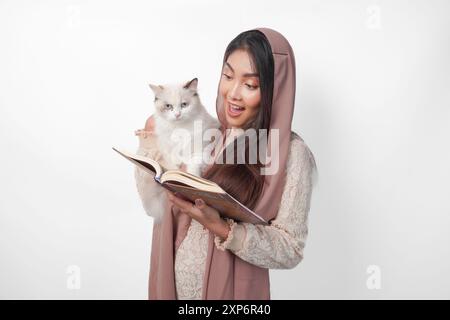 Attraktive junge asiatische muslimische Frau im Schleier-Hijab lächelt, während sie ein weißes Ragdoll-Katzentier umarmt und Al Quran auf der anderen Seite hält Stockfoto