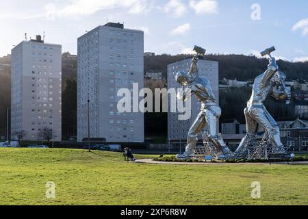 Riesige Schiffsbauer Skulptur „The Skelpies“ des Künstlers John McKenna in Port Glasgow, Inverclyde, Schottland, 2024. Turmblöcke im Hintergrund. Stockfoto