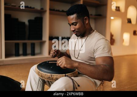 Ruhiger afrikanischer Mann in Trance, der auf Stahlseiltrommel mit Stöcken auf dem Boden im Meditationsraum spielt. Schlagzeuger mit traditioneller Meditation Stockfoto