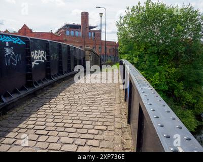 Wigan UK. 28. Juli 2024. Ein kopfsteingepflasterter Pfad führt über einen überbrückten Kanal in einem urbanen Gebiet mit Graffitis-bedeckten Wänden und üppigem Grün. Stockfoto