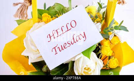 Die Worte HALLO TUESDAY auf einer Visitenkarte in einem Korb mit Blumen, Draufsicht. Das Konzept einer guten Stimmung Stockfoto