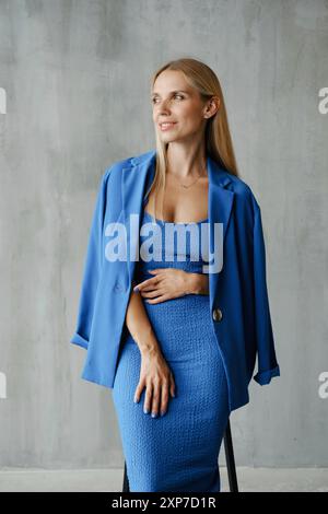 Eine selbstbewusste Frau in einem auffälligen blauen Kleid und Blazer posiert anmutig in einem minimalistischen Ambiente und zeigt ihre Eleganz und Stärke. Stockfoto