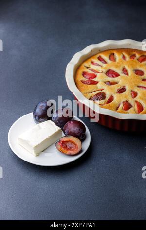 Ein wunderschön gebackener Pflaumenkuchen wird zusammen mit einem Stück Butter und frischen Pflaumen auf einer dunklen Arbeitsplatte serviert. Stockfoto