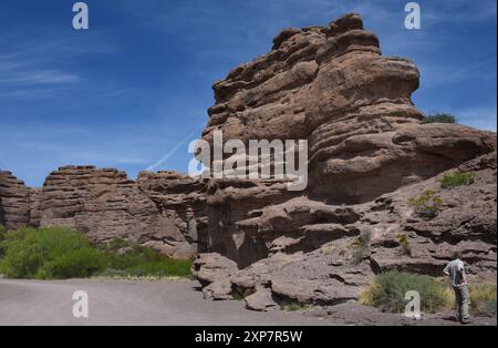 Der Mann steht neben einer großen roten Sandsteinklippe. Er hat braune Khakis und Hemd. Über ihm erhebt sich eine Klippe mit blauem Himmel. Stockfoto