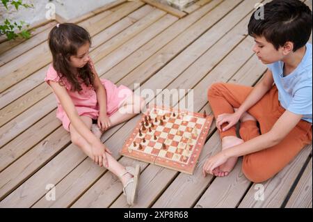 Zwei Kinder sitzen auf einem Holzdeck und spielen ein durchdachtes Schachspiel. Eine unterhaltsame und lehrreiche Outdoor-Aktivität. Stockfoto