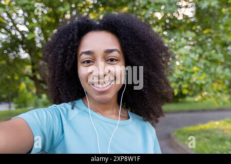 Junge Frau, die im Park im Freien telefoniert, lächelt und mit einem Freund spricht. Sie trägt Ohrhörer und scheint glücklich und engagiert zu sein. Stockfoto