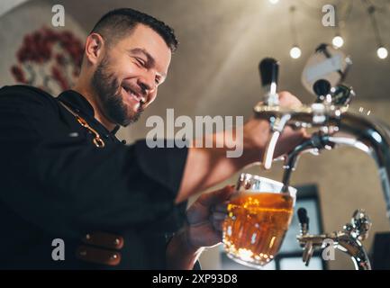 Ein lächelnder, stilvoller Bärenbarmann in schwarzer Uniform mit einer Schürze, der frisches Lagerbier in einen Glasbecher an der Bar zapft. Erfolgreiche Leute, Bierkonzern Stockfoto