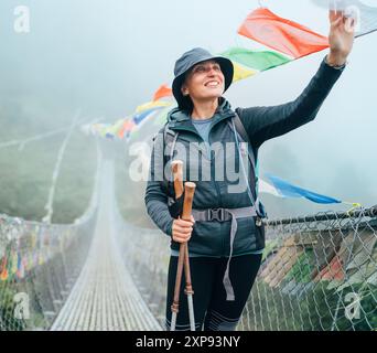 Junge lächelnde Frau überquerte den Canyon über die Hängebrücke und berührte bunte tibetische Gebetsfahnen über der Schlucht. Kletterroute auf dem Gipfel Mera Stockfoto