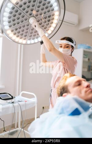 Vertikales Porträt einer Kosmetikerin in Gummihandschuhen, die moderne Lampe über dem Kosmetologiesessel in der Heilklinik des Kursalons andreht. Stockfoto