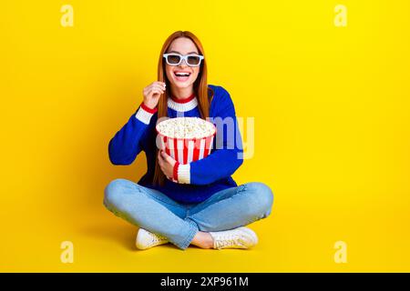 Foto einer jungen Frau in blauem stilvollem Pullover und Jeans, die alleine Popcorn isst und Film auf gelbem Hintergrund sieht Stockfoto