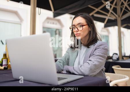 Frau, die außerhalb des Cafés im Freien arbeitet, mit einem Laptop auf der Restaurantterrasse auf der Straße der Stadt sitzt und lernt. Eine engagierte Freiberuflerin, die ein Notebook benutzt Stockfoto
