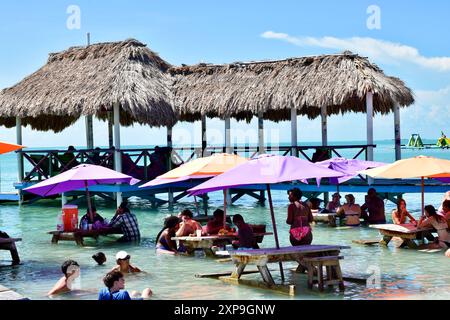 Strandgänger essen und trinken an Tischen im Wasser unter Sonnenschirmen. Im Hintergrund ist eine Palapa zu sehen. Stockfoto