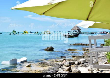 Ein Blick auf Leute, die in einem aufblasbaren Wasserpark am Secret Beach, San Pedro, Belize spielen. Schwimmer, ein Boot, ein Palapa und Sonnenschirme sind ebenfalls zu sehen. Stockfoto