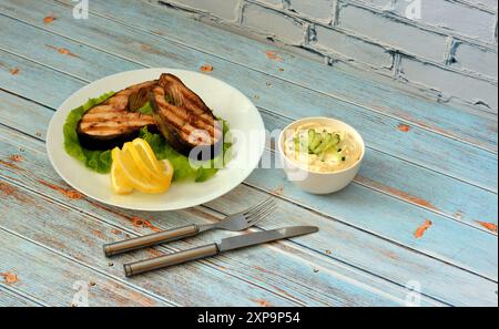 Zwei gegrillte Lachssteaks auf einem Salatblatt mit Zitronenscheiben auf einem hellen Holztisch, neben einer Tasse mit Sauce und Besteck. Nahaufnahme. Stockfoto