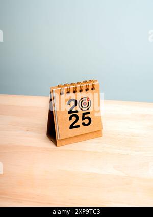 Happy New Year 2025 vertikaler Hintergrund. 2025 Zahlenjahr mit Zieldart-Symbol auf brauner, kleiner Schreibtischkalender, auf Holztisch stehend, minimaler Stolz Stockfoto