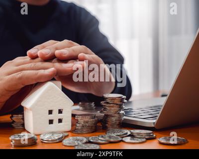 Haus- und Immobilienversicherung, Immobiliendarlehenskonzept. Weißes modernes Hausmodell und viele Münzstapel auf Holztisch, geschützt durch die Hand des Geschäftsmannes oder Stockfoto