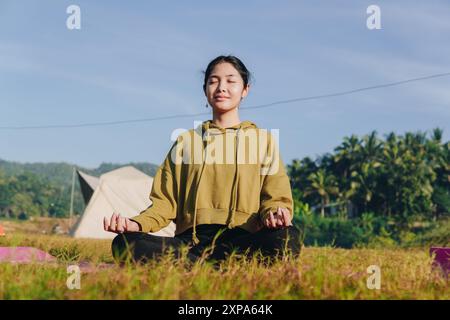 Eine ruhige junge Asiatin sitzt auf einem Feld, trägt einen grünen Hoodie und macht Yoga. Sie ist in einem friedlichen und entspannten Zustand, vielleicht meditiert oder en Stockfoto