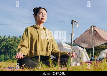 Eine ruhige junge Asiatin sitzt auf einem Feld, trägt einen grünen Hoodie und macht Yoga. Sie ist in einem friedlichen und entspannten Zustand, vielleicht meditiert oder en Stockfoto