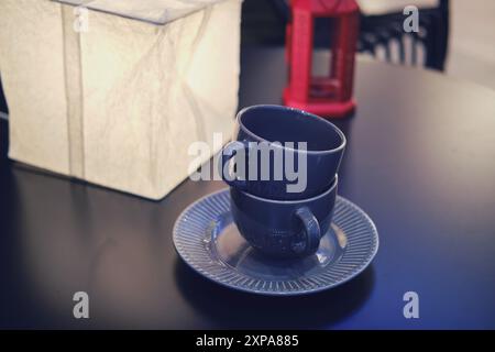 Zwei gestapelte dunkelblaue Teetassen auf einer Untertasse mit weißer Lampe und roter Laterne auf einem schwarzen Tisch. Inneneinrichtung und Tischgeschirr Konzept für Poster und Stockfoto