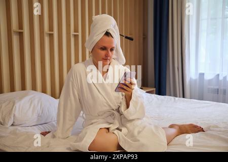 Frau in weißem Bademantel, die mit Telefon auf dem Bett sitzt. Moderne Inneneinrichtung für Wohnkultur und Entspannung Stockfoto