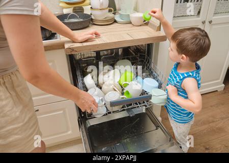 Eine Mutter und ein Kind entladen einen Geschirrspüler in einer Küche. Das Baby hilft Eltern bei der Hausarbeit. Kind im Alter von drei Jahren (dreijähriger Junge) Stockfoto