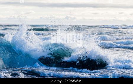Das Meer ist rau und die Wellen stürzen gegen einen Eisberg. Das Wasser ist blau und weiß. Der Himmel ist bewölkt und die Sonne ist nicht sichtbar Stockfoto