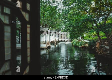 Ein Blick durch ein Holzgitterfenster auf einen traditionellen chinesischen Garten mit Teich, weißem Pavillon und üppigem Grün. Stockfoto