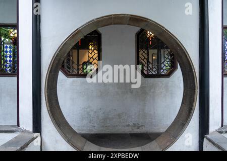 Suzhou, China - 11. Juni 2024 : Eine kreisförmige Öffnung in einer weißen Wand umrahmt zwei achteckige Fenster mit komplizierten Gittern. Stockfoto