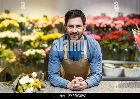 Lächelnder männlicher Blumenhändler in Schürze, der sich auf eine Theke stützt, mit Blumenstrauß umgeben von bunten Blumen im Blumenladen. Person, die wunderschöne Blumenarrangements und Kundenservice in einem blühenden Geschäft präsentiert Stockfoto