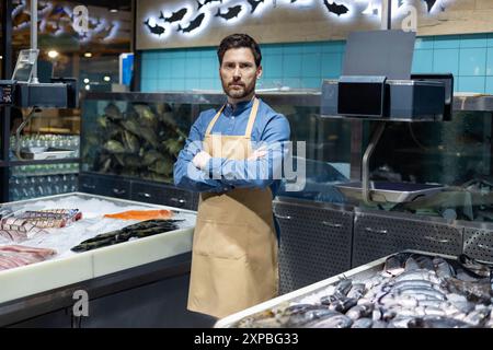 Fischhändler stehen selbstbewusst mit gekreuzten Armen auf dem Meeresfrüchtemarkt, umgeben von frischen Fischvorführungen, die verschiedene Arten von Meeresfrüchten zum Verkauf anbieten. Profi in der Schürze, der in einem Fischhändler arbeitet. Stockfoto