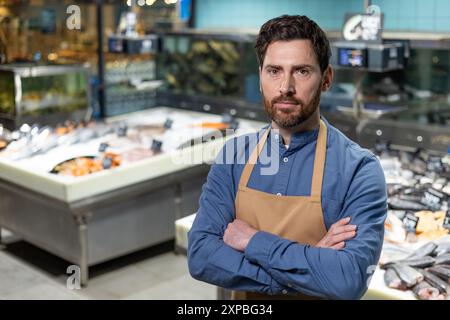 Fischhändler stehen selbstbewusst mit überkreuzten Armen vor frischen Meeresfrüchten im Supermarkt. Mann mit Schürze, der Stolz und Professionalität in seiner Arbeitsumgebung zum Ausdruck bringt. Stockfoto