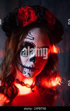 Nahaufnahme einer Frau mit Gesichtsfarbe Day of the Dead und einer Blumenkrone, beleuchtet von dramatischer roter Beleuchtung, die kulturellen und künstlerischen Ausdruck zeigt. Stockfoto