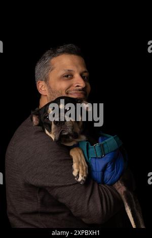 Ein Mann hält einen Hund in den Armen. Der Hund trägt eine blaue Jacke Stockfoto