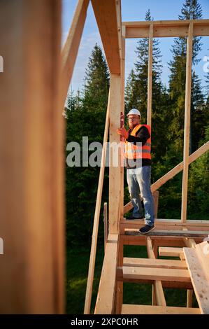Zimmermann baut Holzrahmenhaus in der Nähe des Waldes. Ein bärtiger Mann, der eine Brille trägt, überprüft die Wände mit einer Wasserwaage auf Ebenheit. Er trägt Schutzhelm und orangefarbene Weste. Stockfoto
