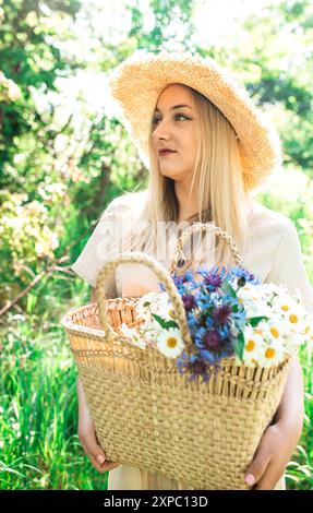 Die schöne Frau hält einen Haufen Gänseblümchen in einem Strohsack auf dem Feld. Sommerlandschaft und junges Mädchen mit Feldblumen. Glückliche Frau in einem Leinenkleid. Stockfoto